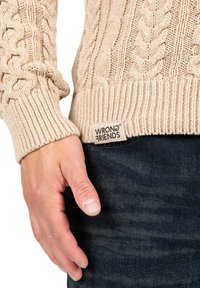 Beige stickad tröja med en strukturerad kabeldesign, ribbade cuffs och en synlig "WRONG FRIENDS"-etikett, bärs över mörk denim.