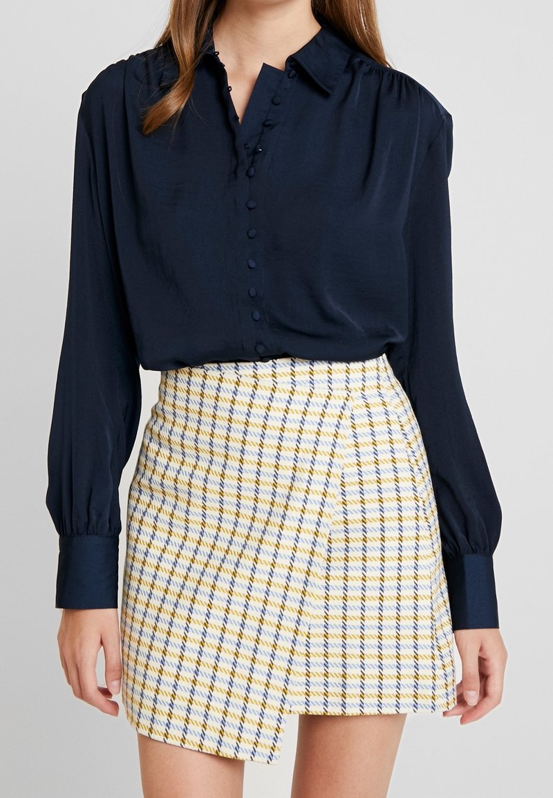 Blusa de manga larga azul marino con detalles de botones y falda corta de cuadros en amarillo, azul y blanco. La falda presenta un diseño envolvente y una tela texturizada.