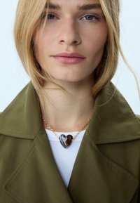 Primo piano di una donna che indossa un cappotto verde oliva e una collana con pendente a forma di cuore marmorizzato su una catena dorata, sopra una maglietta bianca.