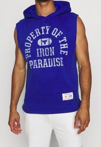 Felpa senza maniche blu con il testo "PROPRIETÀ DEL PARADISO DI FERRO", cappuccio con cordino e etichetta del logo Under Armour in basso.
