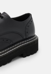 Chaussure en cuir noir avec un design brogue, épaisse semelle plateforme noire et accents de couture blanche. Présente une texture lisse et une forme arrondie.