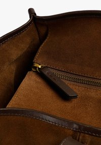 Interior de un bolso de ante marrón con un bolsillo con cremallera forrado en tela verde olivo, herrajes dorados y detalles en cuero marrón.