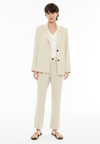 Traje beige a rayas con una chaqueta a medida que cuenta con dos botones en el frente y pantalones a juego. Lleva puesto un top blanco de cuello en V, combinado con sandalias.