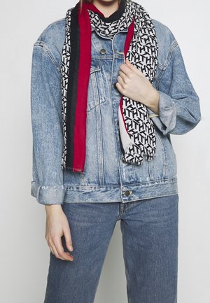 Personne portant une veste en denim bleu clair retroussée et un jean, avec une écharpe noire, blanche et rouge à motifs enroulée autour du cou.