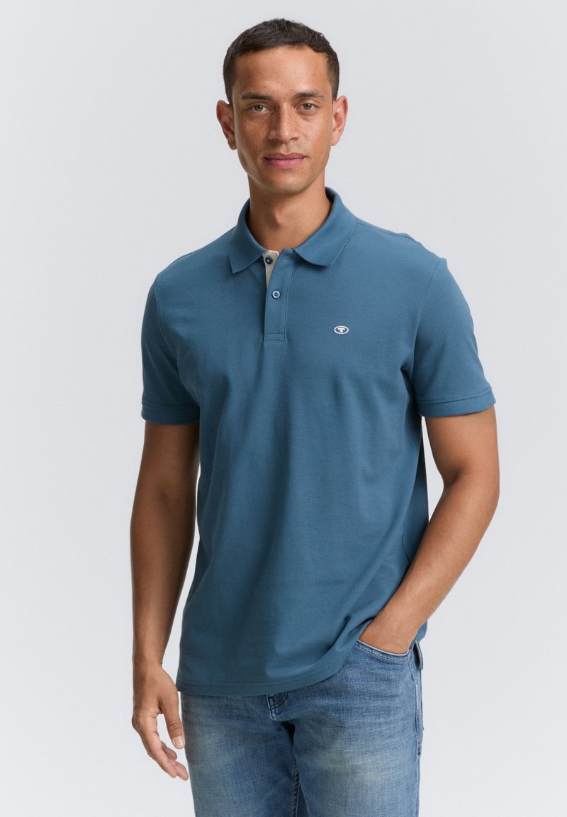 Camisa polo de algodón azul con un cuello beige en contraste y dos botones, que presenta un pequeño logo en el lado izquierdo del pecho. Se lleva con jeans de color azul claro.