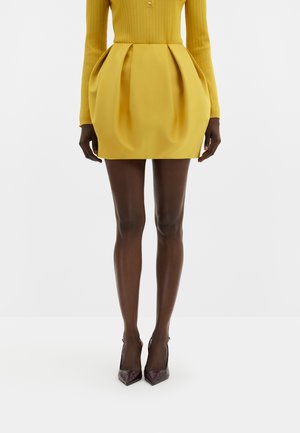Femme portant une mini-jupe plissée jaune, un haut côtelé à manches longues assorti et des escarpins à bouts pointus foncés sur fond blanc.