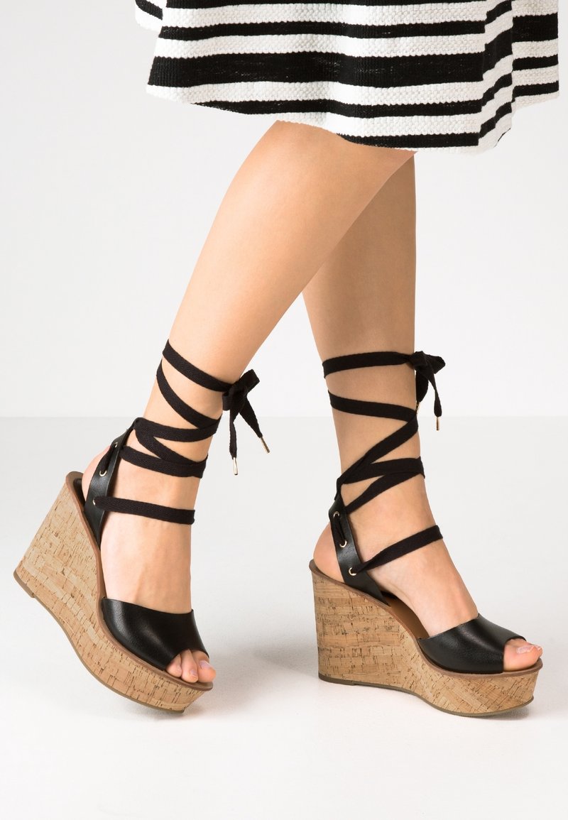 Sandales compensées en cuir noir avec talon en liège, brides de cheville enroulées et nouées, et design à bout ouvert. Contraste de textures entre les matériaux.
