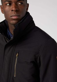 Czarna kurtka z wysokim kołnierzem, z kieszenią na zamek błyskawiczny na klatce piersiowej i teksturowanym materiałem. Złote elementy zamka kontrastują z całością.