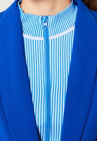 Blå blazer över en blå och vit randig topp. Toppens framsida har en dragkedja och ribbad textur runt halsringningen.