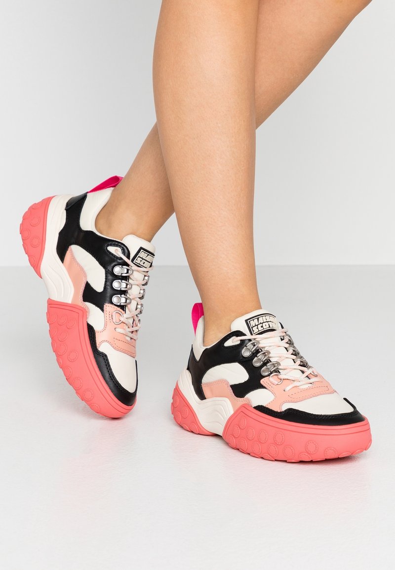 Jambes portant des baskets à semelles épaisses avec des semelles roses, des panneaux noirs, blancs et pêche, et des lacets roses sur un fond blanc uni.