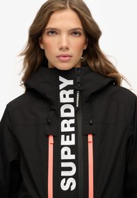 Czarna kurtka z wysokim kołnierzem, z wyraźnym białym logo "SUPERDRY" oraz pomarańczowymi uchwytami zamków; wykonana z gładkiego, trwałego materiału.