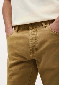 Pantalons en coton kaki à coupe droite, dotés de poches latérales, d'une fermeture à bouton et d'une texture subtile. Portés avec une chemise de couleur claire.