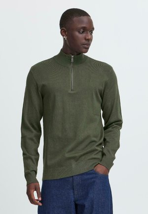 Olivenfarget genser med glidelås og ribbestruktur, med lange ermer og en tettsittende design. Akkompagnert av en kort sølvglidelås ved kragen.