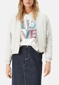 Crème bomberjack met ribbelaccenten, gedragen over een witte hoodie met kleurrijke "LOVE" tekst, gecombineerd met een donkere spijkerrok.