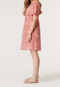 Röd- och vitmönstrad klänning med korta ärmar, V-ringning och markerad midja, kombinerad med ljusa remsandaler.