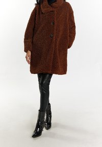 Brun, struktureret teddyfrakke med sjalskrave, tre sorte knapper og oversized pasform, parret med tætsiddende sorte bukser i kunstlæder og støvler.