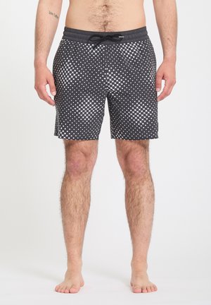 Torso et jambes d'un homme portant un short de bain noir avec un motif de points et carrés blancs, taille élastique avec cordon noir, debout pieds nus.