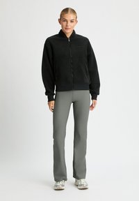 Giacca zip-up nera in pile con maniche lunghe, polsini ed elastico in vita. Abbinata a pantaloni grigi slim-fit e scarpe da ginnastica.