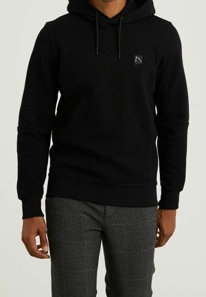 Homme portant un sweat à capuche noir avec cordons et un pantalon à carreaux gris, debout devant un fond clair uni.