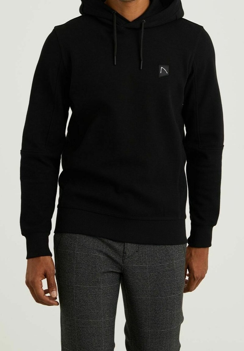 Homme portant un sweat à capuche noir avec cordons et un pantalon à carreaux gris, debout devant un fond clair uni.