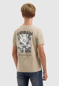Jong persoon met de rug naar ons toegekeerd, gekleed in een beige T-shirt met een zwart-wit stedelijk thema grafisch ontwerp en de tekst "URBAN JUNGLE" op de achterkant.