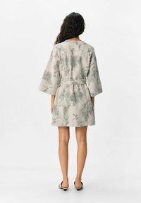 Donna con lunghi capelli ricci che indossa un vestito beige a fantasia floreale con cintura e sandali con tacco bianchi e neri, in piedi di spalle.