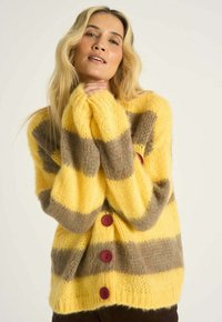 Žena s dlhými blond vlasmi, ktorá má na sebe žlto-hnedý pruhovaný plyšový sveter s červenými gombíkmi, ruky zložené pri tvári, neutrálny pozadie.