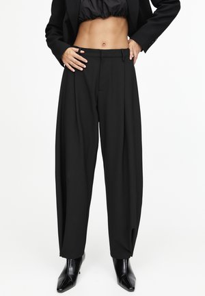 Black high-waist wide-leg trousers worn with black ankle boots and a cropped black jacket, hands resting on hips, against a white background.