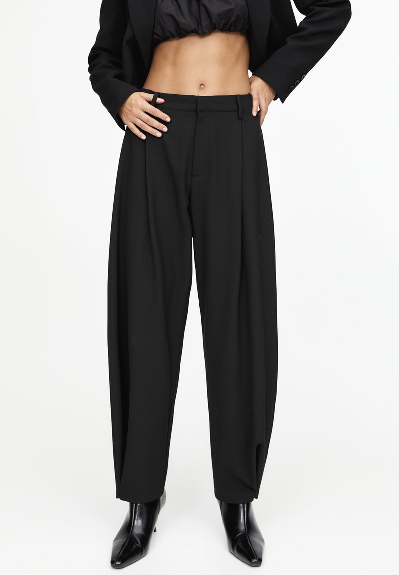 Pantalon noir taille haute à jambes larges, porté avec des bottines noires et une veste courte noire, mains sur les hanches, sur fond blanc.