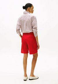 Shorts rouges avec une poche arrière, associés à une chemise à rayures à boutons, en tissu léger. Le modèle porte des mocassins blancs.