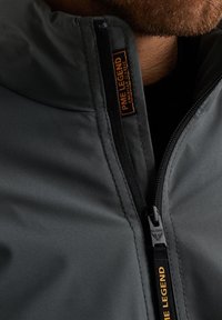 Chaqueta de nailon gris con un alto cuello, cierre de media cremallera y una etiqueta de la marca "PME Legend" en naranja. Textura suave con costuras mínimas.