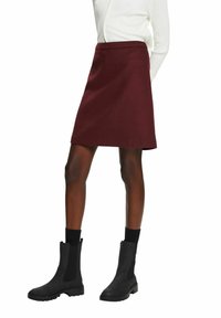 Femme portant une jupe trapèze bordeaux associée à des bottines noires et des chaussettes noires, debout devant un fond blanc.