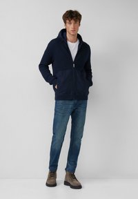 Navy hooded zip-up sweater with ribbed texture and solid lower panel, paired with denim jeans and brown lace-up boots.