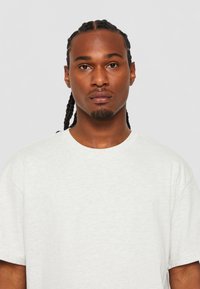 Lichtgrijs, t-shirt met korte mouwen en een ronde halslijn. De stof lijkt zacht met een gladde textuur. Het model heeft gevlochten haar en een neutrale uitdrukking.