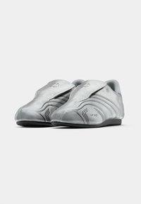 Ezüst sportcipők elegáns kialakítással, ívelt sziluettel, sima textúrával és tépőzáras pánttal, diszkrét fekete részletekkel és márkajelzéssel.