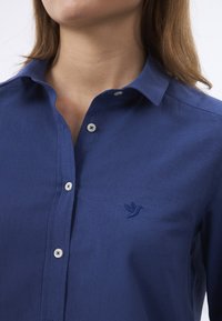 Marineblaues Hemd mit Kragen, ausgestattet mit weißen Knöpfen und einem gestickten Vogel-Logo auf der Brust. Glattes Material mit feiner Textur.