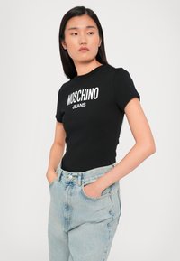 Juoda trumparankovė marškinėlių su balta tekstu "MOSCHINO JEANS", derinama su aukštu liemeniu šviesiai mėlynomis džinsinėmis kelnėmis. Lygus medvilnės audinys.