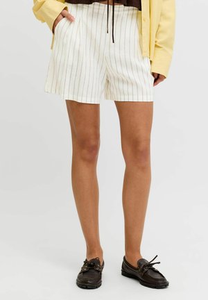 Le mannequin porte un short crème à fines rayures, une chemise jaune et des mocassins marron foncé.