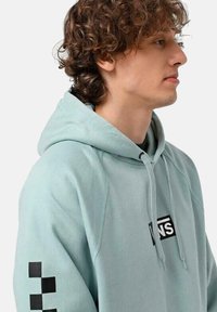 Sudadera con capucha de color azul claro, con un logo en la parte frontal y un patrón de cuadros negros en la manga izquierda. Tejido suave, corte relajado y capucha con cordón.