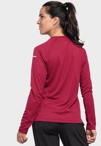 Rotes Langarm-Performanceshirt mit strukturiertem Muster, ausgestattet mit engen Ärmeln und Rundhalsausschnitt, kombiniert mit schwarzen Hosen.