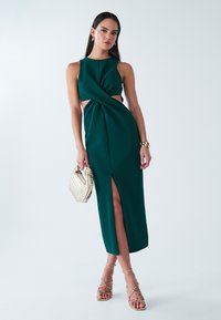 Obleka brez rokavov smaragdno zelene barve s prepletenim detajlom spredaj, stranskimi izrezi in visokim razporkom ob stegnih; kombinirana z zlatimi sandali z trakovi in pleteno torbico.