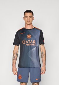 Svart och mörkblå sporttröja med orange detaljer, med Qatar Airways-logotyp och vågig grafisk design; matchad med blå shorts.