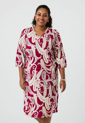 Mujer sonriente, vestida con un vestido hasta la rodilla y manga tres cuartos, con un estampado de paisley en rojo, blanco y beige, de pie contra un fondo claro y liso.