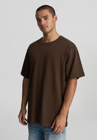 T-shirt in cotone marrone con scollo rotondo, maniche corte e vestibilità rilassata. Il tessuto ha una texture liscia. Indossato con jeans azzurri chiari.