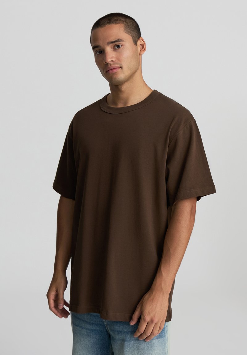 T-shirt in cotone marrone con scollo rotondo, maniche corte e vestibilità rilassata. Il tessuto ha una texture liscia. Indossato con jeans azzurri chiari.