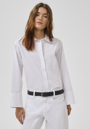 Woman with long brown hair wearing a white button-up shirt tucked into white pants with a black belt against a plain background.