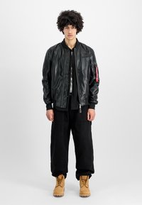 Μαύρο δερμάτινο μπουφάν bomber με ριγωτό γιακά, φερμουάρ μπροστά και κόκκινη ετικέτα στο μανίκι. Συνδυασμένο με φαρδιά μαύρα παντελόνια και κίτρινες μπότες.