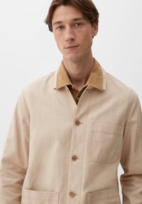 Beżowa bawełniana kurtka o teksturowanym wykończeniu, z brązowym kołnierzem, z przodu zapinana na guziki i z kieszenią na piersi, zaprojektowana w luźnym fasonie.