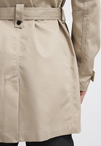 Détail de la taille d'un trench-coat beige avec passants de ceinture fermé par un bouton noir, montrant une partie d'un bras et un pantalon noir.
