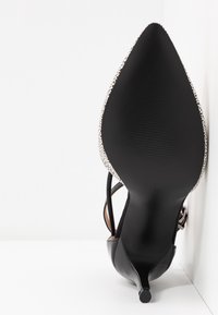 Chaussure à talon haut noire à bout pointu avec un talon kitten fin et des bords blancs tachetés, affichée semelle vers le haut sur fond blanc.